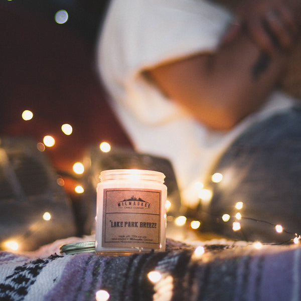 6 ounce Lake Park Breeze soy wax candle on a blanket with fairy lights surrounding it. There's a blurry body sitting down in the background.