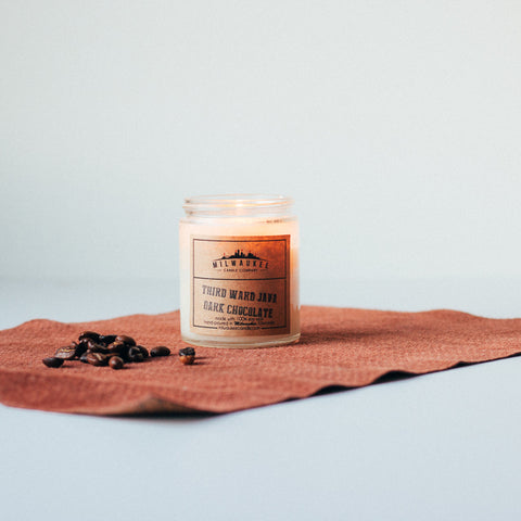 Lit 6 ounce Third Ward Java Dark Chocolate candle sitting atop a piece of brown suede fabric with a pile of coffee beans next to it.